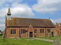 A small, simple, red sandstone church seen from the south. On its left is a small spirelet with a clock, and a small porch protrudes from the south wall