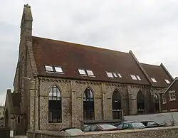 Side view of a tall, narrow flint building with a steeply-pitched red-tiled roof in which several skylights have been inserted at irregular intervals. Four identical lancet window openings with modern windows, separated by buttresses, dominate the near side; an identical window is visible on the recessed side wall behind it. A stone bell-tower rises from the left-hand side, and below it a porch projects forward a short way.