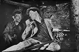 Aleksei Stakhanov and another man at work in a Soviet coal mine. Stakhanov, while holding a drill, is seated at the coal face, his head turned to speak to his colleague.