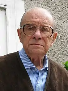 A bust photograph of an older white man; he has little hair, is wearing large glasses and looking into the camera unsmiling.