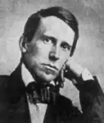 A man in his 20's rests his hand on his chin, while wearing a formal jacket and a bow tie