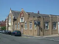 Three-quarter view of a brownish building on a corner site, with cars parked in front of its main façade.  This has a single storey with two taller gabled sections, the larger of which (furthest away in the picture) has a short tower-style structure next to it.  A black and white sign hanging from the corner is headed "Stoneham Road Baptist Church".