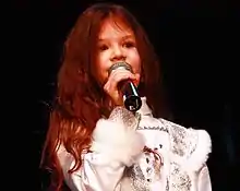 A young woman singing into a microphone in front of a black background, wearing a white blouse.