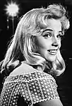 Close-up black-and-white portrait photo of a smiling young woman with long blonde hair in a studio, brightly illuminated by set lights