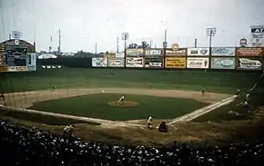 A green baseball field with a sloping right field wall surrounded by colorful billboard advertisements.