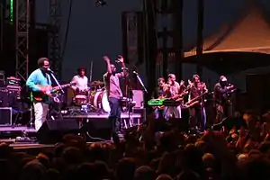 All-male group on stage containing a guitarist, a drummer, a vocalist, a keyboardist, and three members of a horn section. Some of the crowd present in the foreground