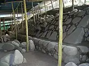 Excavated area with a flat dusty area to the left with some rubble and with steeply sloping well-dressed stonework to the right sat on top of a low vertical stonework base. A ramshackle roof protecting the excavations is supported by wooden poles.