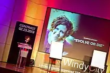 Shannon Downey stands before a podium on a stage. Large text on the stage reads "TEDx Windy City". Also on the stage is a projector screen with a presentation slide. On the slide is a picture of Downey and the title "Evolve or Die".