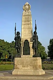 A cream-coloured stone obelisk bearing names rises into a blue sky. Above its base plinth is a large, black plaque bearing the words "Their names liveth for evermore" and "Teen Murti". On either side on its plinth is a statue of a turbaned man in military uniform holding aloft a weapon with a flag at its top. Behind the obelisk is a garden with pink flowers and trees, and a road recedes into the distance.