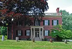 A six-bay-wide brick house with green window shutters, a balustrade on the roof and an entrance porch with round fluted Ionic columns. Along the front lawn, which rises slightly, is a line of five tree stumps with bark removed. There is a large tree on the left partially shading the house.