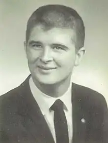 A monochromatic bust photo of a suited young white man with a buzz cut; he is smiling and facing the camera, looking to its left