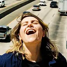 A photograph of Alex Lahey with her head tilted back, eyes closed and mouth open in front of cars on a highway