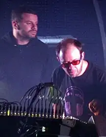 A dark image of two men in the back playing the synthesizers.