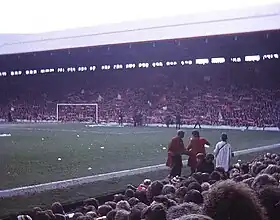 Photograph of a stand full of people. In front of the stand is a field and a goal.