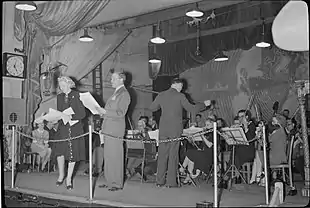 Photograph taken at a live performance, featuring two performers at the front of the stage backed by the band and conductor