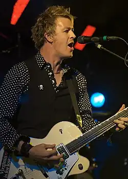 Cheney is singing at a microphone while playing his guitar. He is shown in right profile, he wears a dark shirt with white polka dots, covered by a sleeveless vest. Some stage lights are visible behind him.