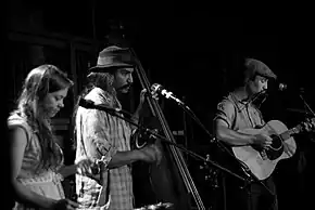 A black-and-white image of three musicians (a woman and two men) playing instruments.