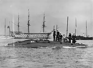 A group of sailors standing around an access hatch atop a partially submerged vessel with sailing ships behind them