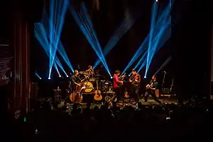 Five male musicians perform on stage in front of a standing audience, behind them a dozen lights project blue lines upward.