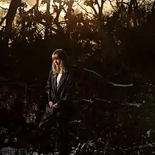 Tamara Lindeman stands in a forest, staring off to the left. The forest is dimly lit, though she is illuminated, with a golden hour sunset shining through the row of silhouetted trees behind her.