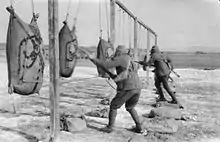 Black and white photograph of three men wearing military uniforms and large sack-like gas masks over their head using bayonets fixed on the end of rifles to attack hessian sacks hanging from a frame