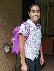 A girl in a school uniform wearing a purple backpack stands in front of a house.