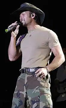 A man wearing a cowboy hat, light T-shirt and camouflage pants singing into a microphone