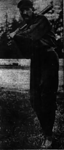 A man with a large mustache wearing a dark baseball uniform and cap stands poised to swing his bat.