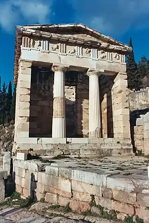 A partially ruined marble building with a porch with 2 columns supporting a pediment and an open doorway beyond