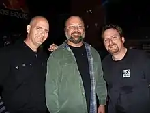 three middle-aged men stand together facing the camera and smiling