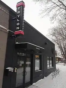 A black storefront with a vertical sign reading "TRYLON" projecting from high on the wall. There is snow on the ground and a set of three bike racks in front of the cinema.