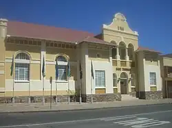 A yellow and white neo-classicist building in bright sunlight. The roof of corrugated iron sheets is painted dark red, on the main gable the inscriptions read "A.D.1913" and "SADC Tribunal". Seven flagpoles with national flags of countries from the Southern African Development Community are mounted onto the pavement in front of the building.