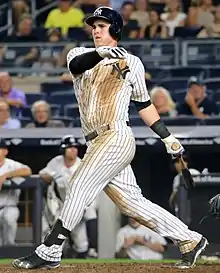 A man, wearing a dirt-stained Yankees pinstripe uniform and batting helmet with the interlocking "NY" logo, follows through after swinging at a pitch.