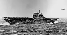 Black and white photo of a flat-decked ship at sea. It is carrying aircraft on the rear of its deck, and one plane is flying immediately in front of the ship.