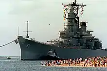 A large gray ship moves toward the camera and slightly to the left. On the right, a number of people on a beach are observing the ship as she moves in to pass them with the aid of a tugboat.