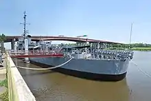 A gray ship in the water, seen from starboard bow, with a gangplank connecting it to land. There is a bridge in the background.