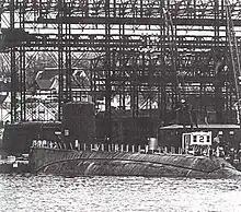 An incomplete submarine lays a rest; tall steel gantries and a dock dominate the background.