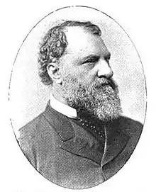 Profile of a white man with curly hair, a receding hairline, and a full beard, wearing a suit coat over a shirt and tie.