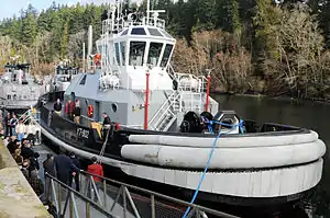 Tugboat Valiant is moored to a pier. A man at a lectern aboard the tugs peaks to an audience on the pier.