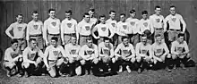 A group of 23 young men arranged in two rows—thirteen standing in back and ten squatting in front—in rugby uniform: white sweaters emblazoned with vertically-striped shields, black belts, and light colored shorts.