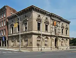 A light tan two-story building with ornamented windows and a colonnade in the center of one side seen from across an intersection, with yellow traffic lights in front.