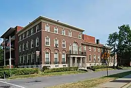 A three-story brick building with a flat roof and colonnaded entrance portico, seen from across the street. To its right is a long two-story wing, and a more modern wing is partially visible to its rear.