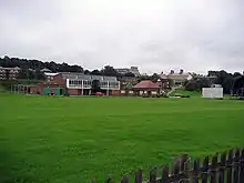 A cricket pavilion  joined to a more modern building behind green playing fields, with the sight screens of a cricket pitch to the right