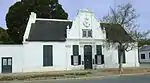 5 bay large saddle-roofed house with covered front and end gables and long verandah. Saddle roof with covered front gable with bargeboarding and semicircular loft window. Walls are rusticated and have eyebrow mouldings. Verandah with hipped ogee roof o Architectural style: Victorianized Cape Dutch style.. Type of site: House Current use: Residential.