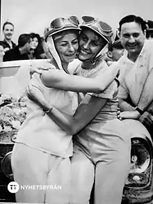 Two white women in helmets, hugging, outside a car.