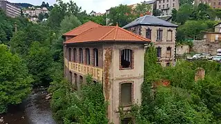 Photo of the Seychalles bridge plant, showing how the entire narrow valley space was used. The 3-storey building is very narrow, angled to match the shape of the riverbank. The roof has been redone, but the floors are still apparently empty and all the door frames are missing. The word "Orfèvrerie" is still clearly visible on the façade.