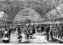 The interior of the arena is shown without ice. Around the arena are tables of plants. Dozens of persons are walking around or inspecting the plants.