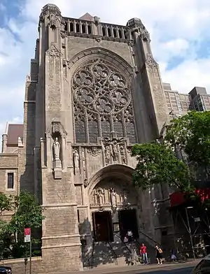 A brown stone tower with rosette window and other decorative touches rising up from a street with trees