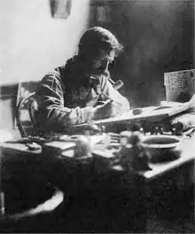 Black-and-white photograph of a man with a large mustache and a pipe in his mouth, at work, seated behind a cluttered desk on which a drawing board is propped at a slight angle.