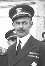 Head and shoulders of a white man with a handlebar mustache, wearing a peaked cap and a dark jacket with two rows of ribbon bars on the left breast. Several other men in similar clothing can be seen behind him.